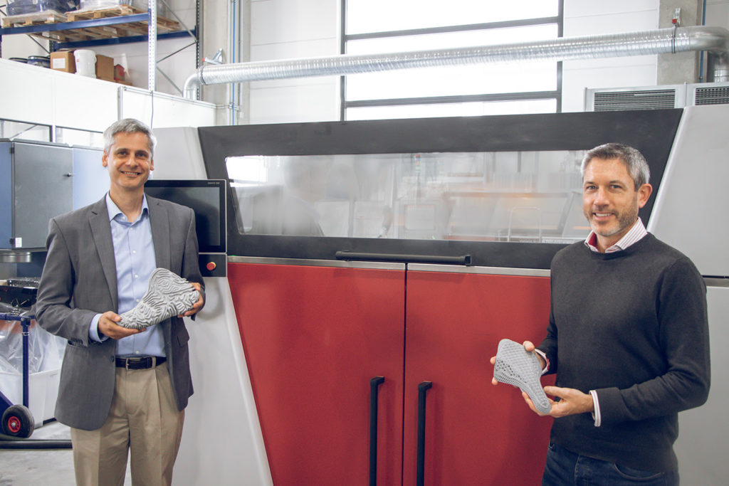 Geoff Gardner, Innovation Director Additive Manufacturing at Covestro (left) and James Reeves, Global Director for Polymer Printing at voxeljet (right) in front of the VX1000 HSS with parts printed with TPU from Covestro